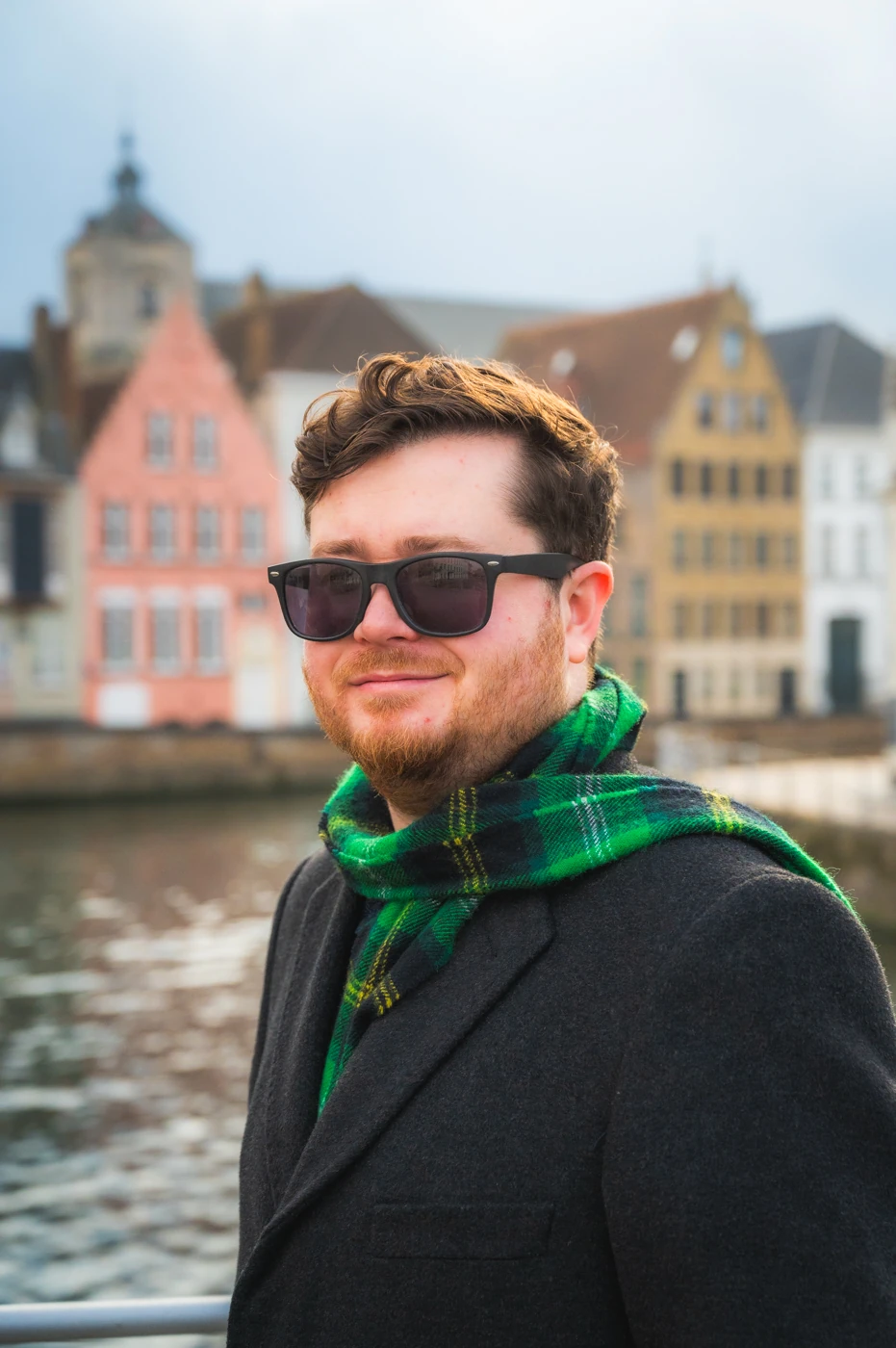 Man with sunglasses, a green scarf and a dark overcoat smiling next to the canal in Bruges, Belgium