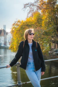 Woman with sunglasses leaning on a railing beside a canal, autumn trees in the background.